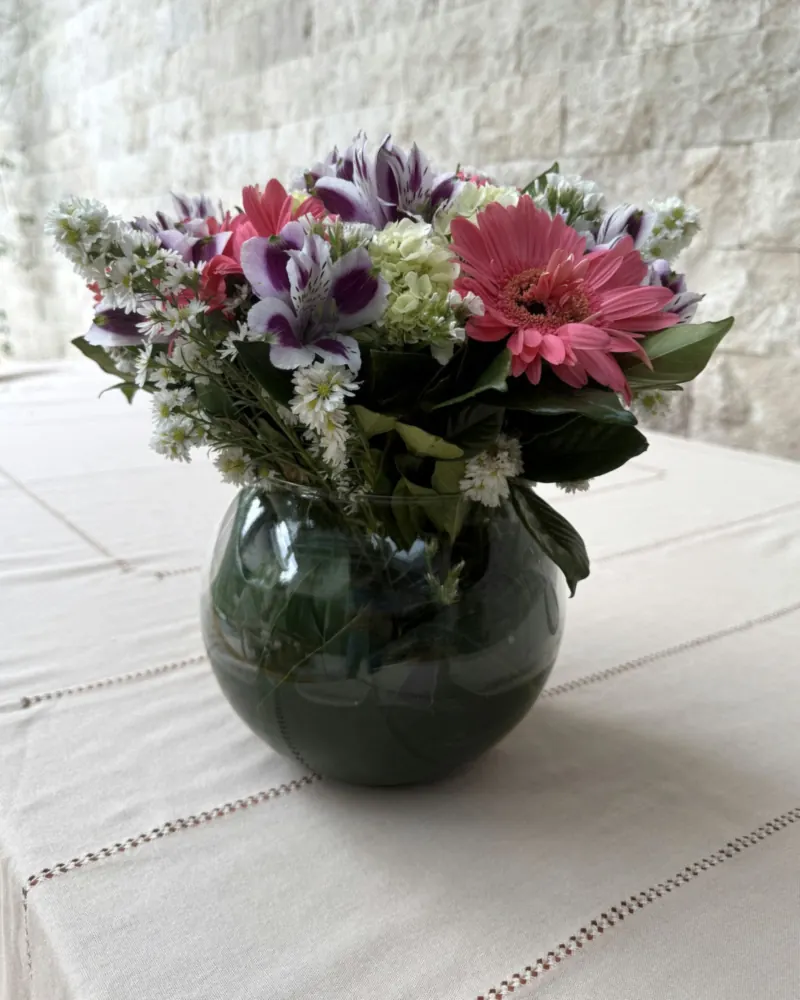 Arranjo de flores visto de frente: gérbera pink aberta, astromélias brancas e roxas e hortênsia verde-clara
