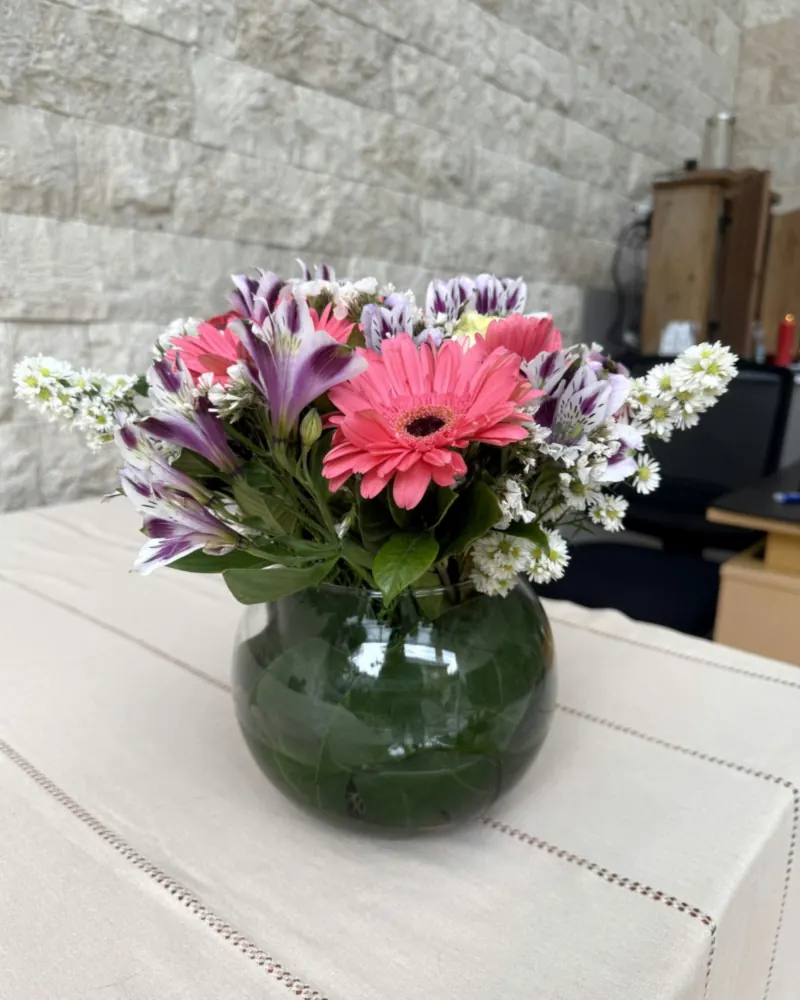 Arranjo de flores naturais em vaso redondo transparente sobre mesa com toalha clara e parede de pedra ao fundo