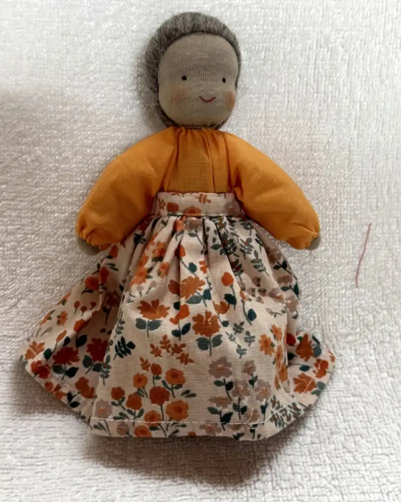 Boneca avó de frente sobre manta branca, com cabelo cinza, blusa laranja e saia longa creme estampada de flores