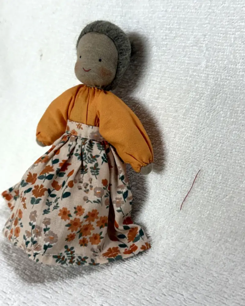 Boneca avó vista em ângulo, com bochechas rosadas, blusa laranja e saia de flores laranja e verde-petróleo