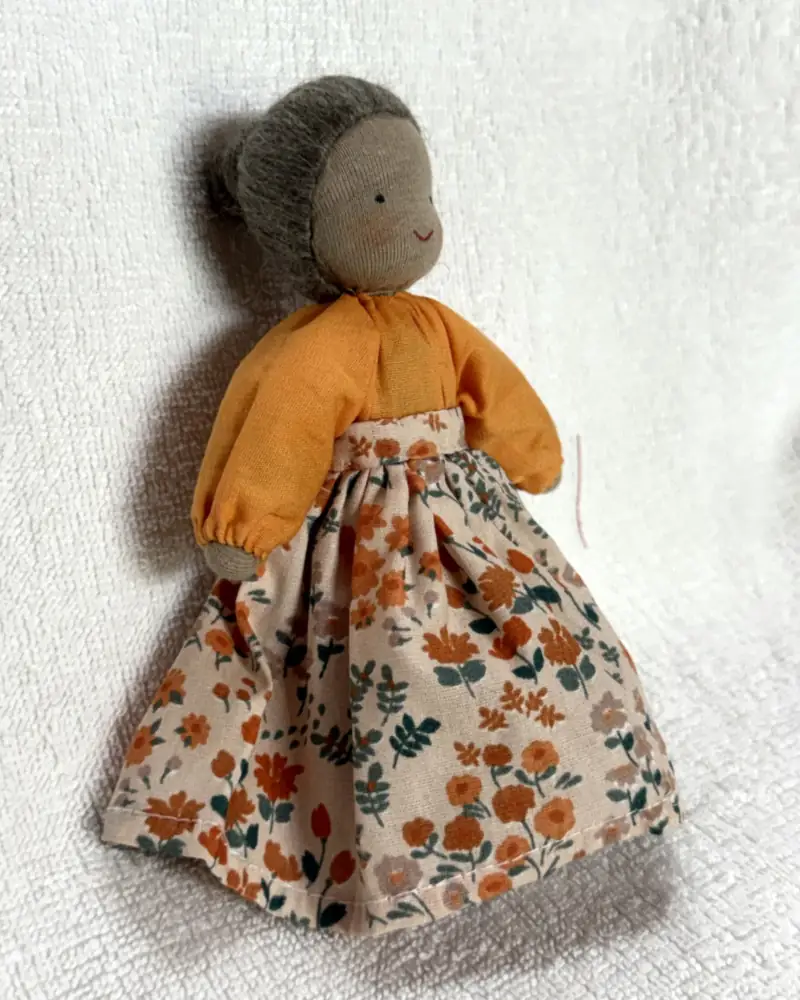 Boneca avó de perfil, mostrando o coque do cabelo cinza, as mangas bufantes laranja e a saia longa florida