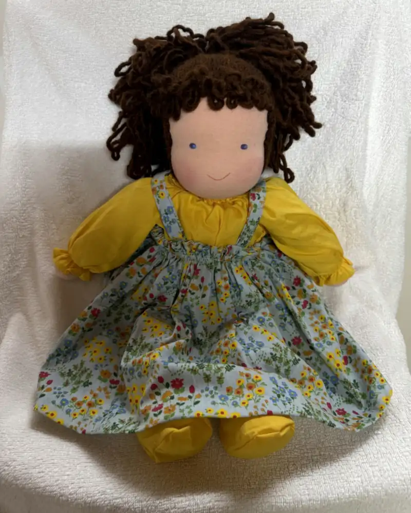 Boneca de cachos castanho-escuros com franja, blusa amarela e jardineira azul-clara florida, sentada de frente