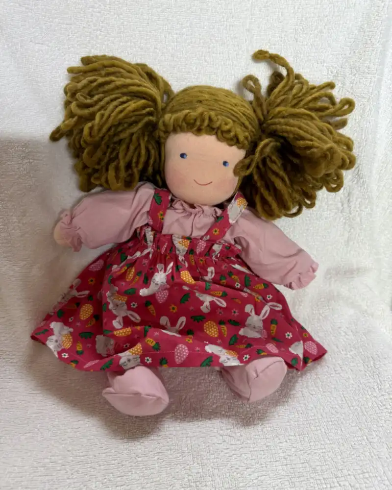 Boneca de marias-chiquinhas, blusa rosa e jardineira pink com coelhinhos, cenouras e ovinhos, sentada de frente