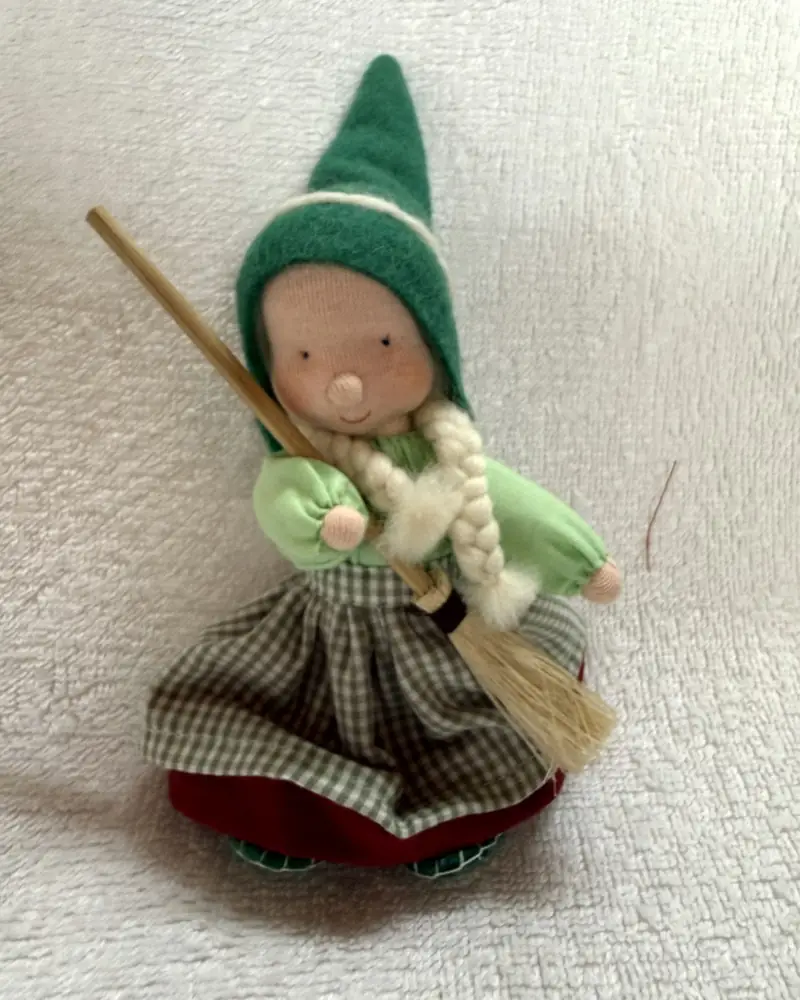 Bruxinha Sapeca de frente sobre manta branca, com gorro pontudo verde, tranças creme e vassourinha de palha