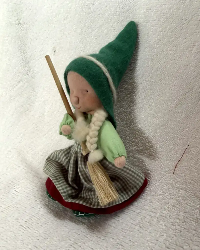 Bruxinha de gorro verde vista em ângulo, segurando a vassourinha, com blusa verde-clara e saia xadrez verde e creme