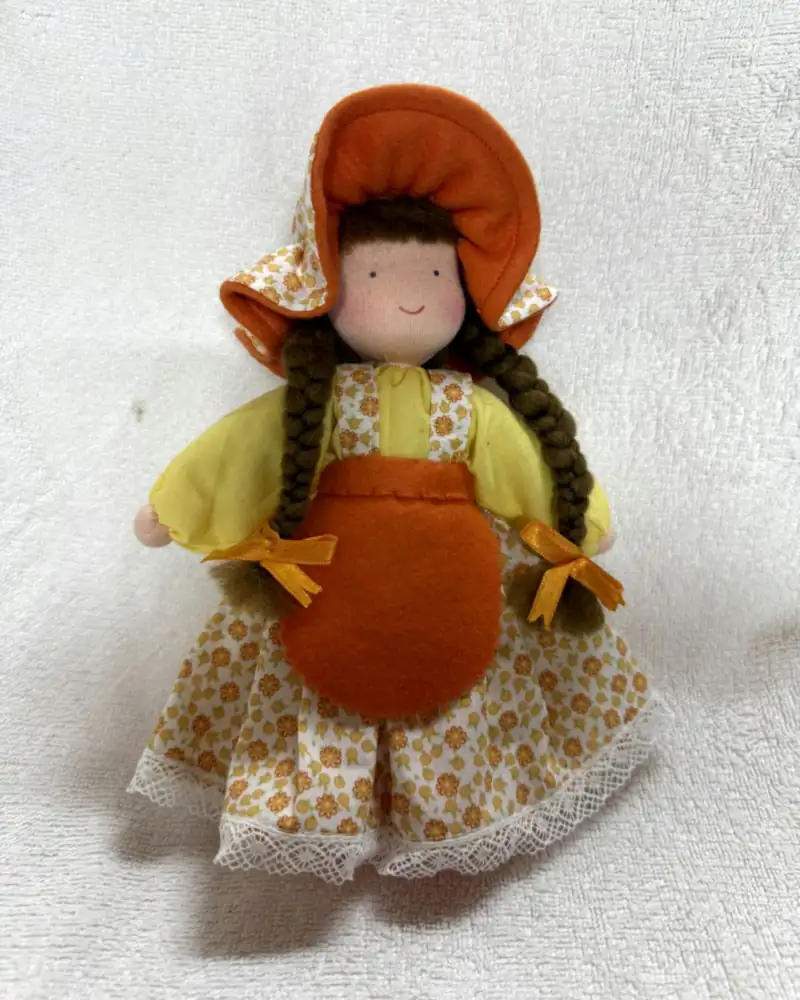 Boneca Chapeuzinho de frente sobre manta branca, com touca laranja, tranças castanhas compridas e avental laranja