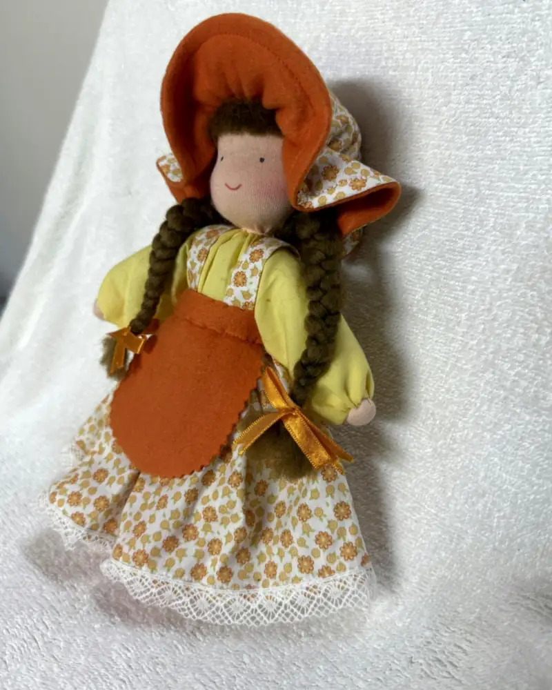 Chapeuzinho vista em ângulo, com blusa amarela, laços de cetim laranja nas tranças e vestido branco de florzinhas
