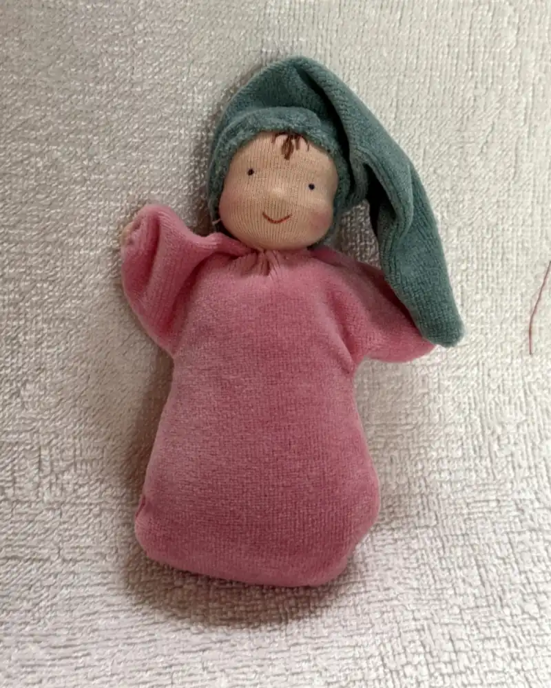 Cheirinho, boneca de pelúcia rosa com gorro pontudo verde-água e cabelinho castanho, de frente sobre manta clara