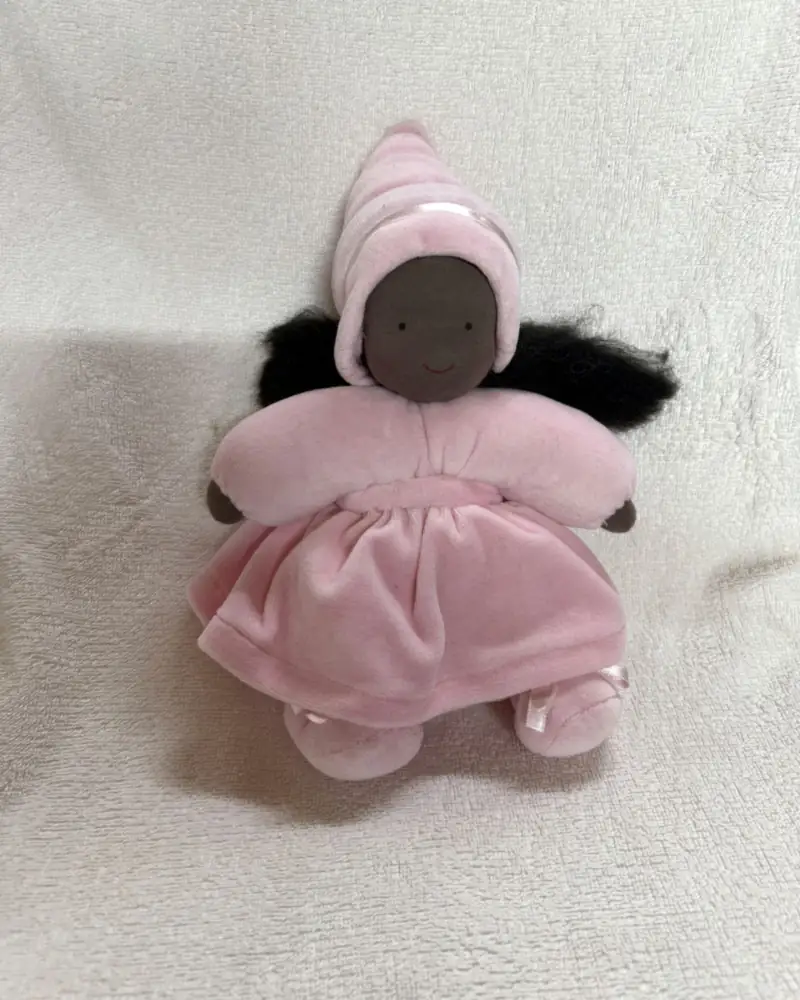 Fadinha de pele negra e cabelo crespo preto, com gorro pontudo e vestido rosa aveludado, sentada sobre manta branca