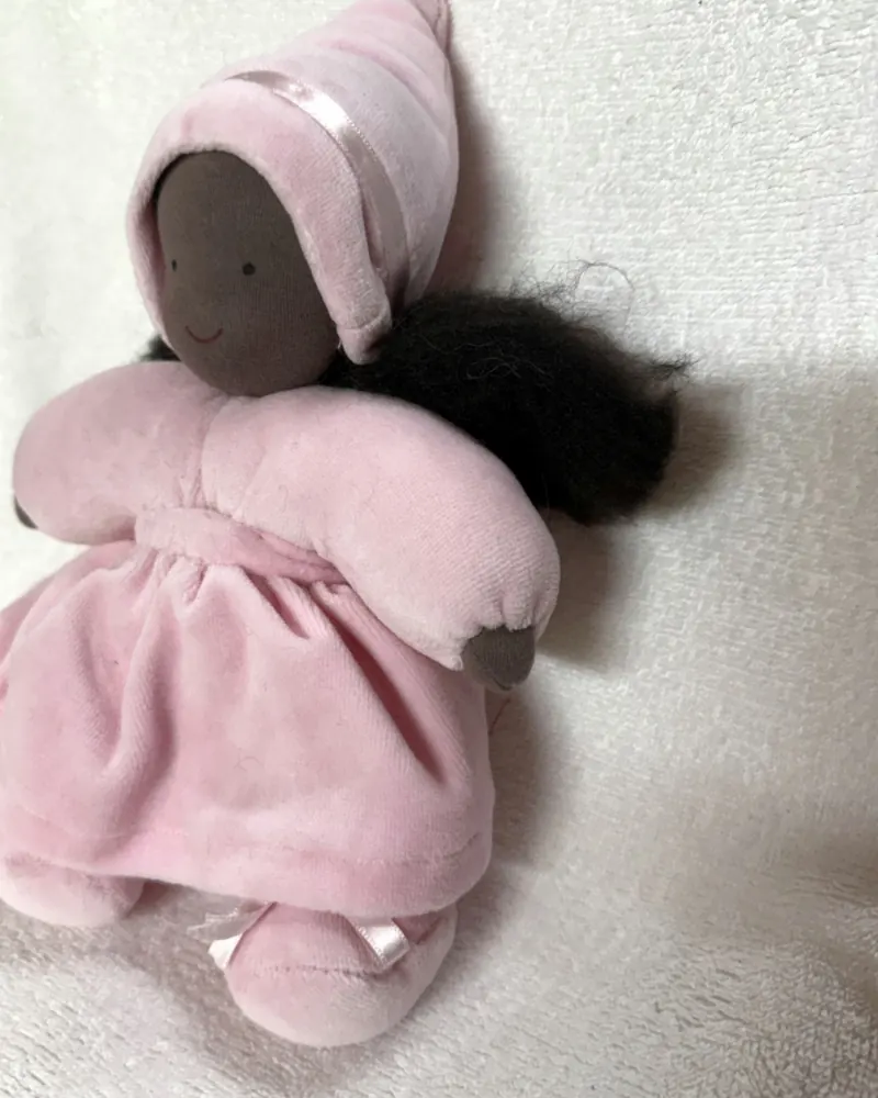 Fadinha de gorro rosa vista de perto, com rosto de pele negra, cabelo crespo preto e sapatinhos rosa de lacinho