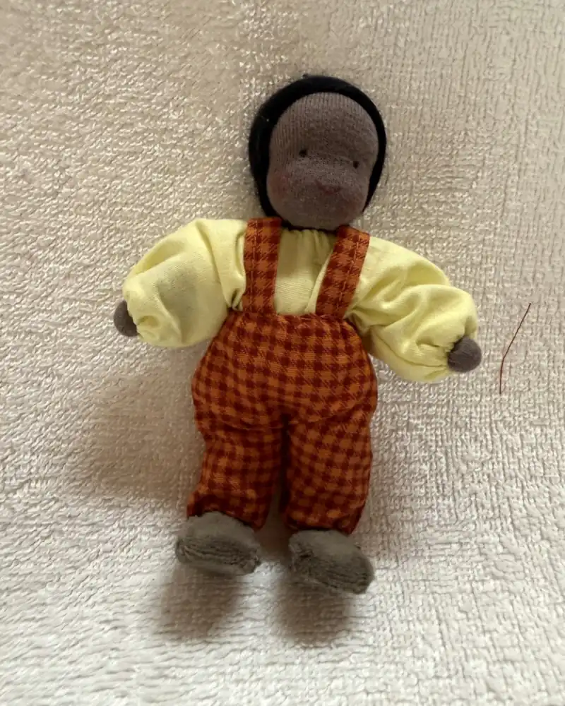 Boneco de pele negra e cabelo preto, com blusa amarelo-clara e macacão xadrez laranja e vinho, deitado de frente