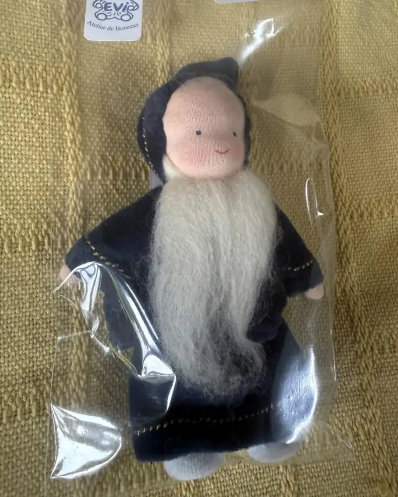 Merlin visto de perto no saquinho, com bochechas rosadas, capuz pontudo e barba branca e cheia sobre o peito