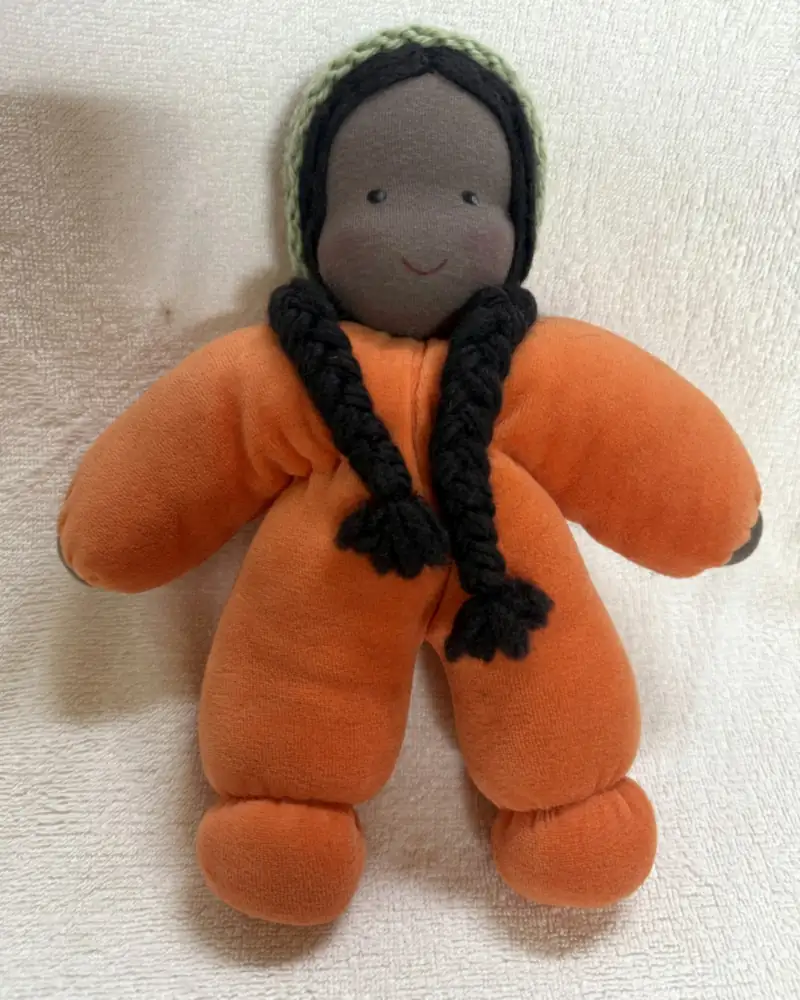 Molinha Laranja, boneca de pele negra, com tranças pretas, touca de tricô verde-clara e corpo de pelúcia laranja