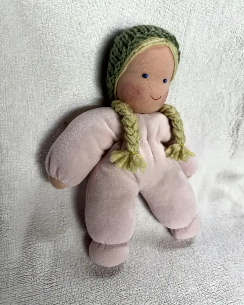Molinha Rosa sentada, com corpo de pelúcia rosa-clara, tranças loiras e touca de tricô verde