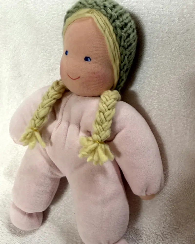 Rosto da Molinha Rosa de perto, com olhos azuis, bochechas coradas e a touca de tricô verde
