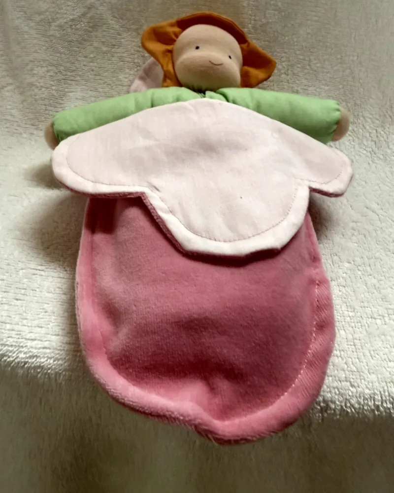 Boneca de colo com touca de pétalas laranja, braços verdes, gola de pétala rosa-clara e corpo de pelúcia rosa