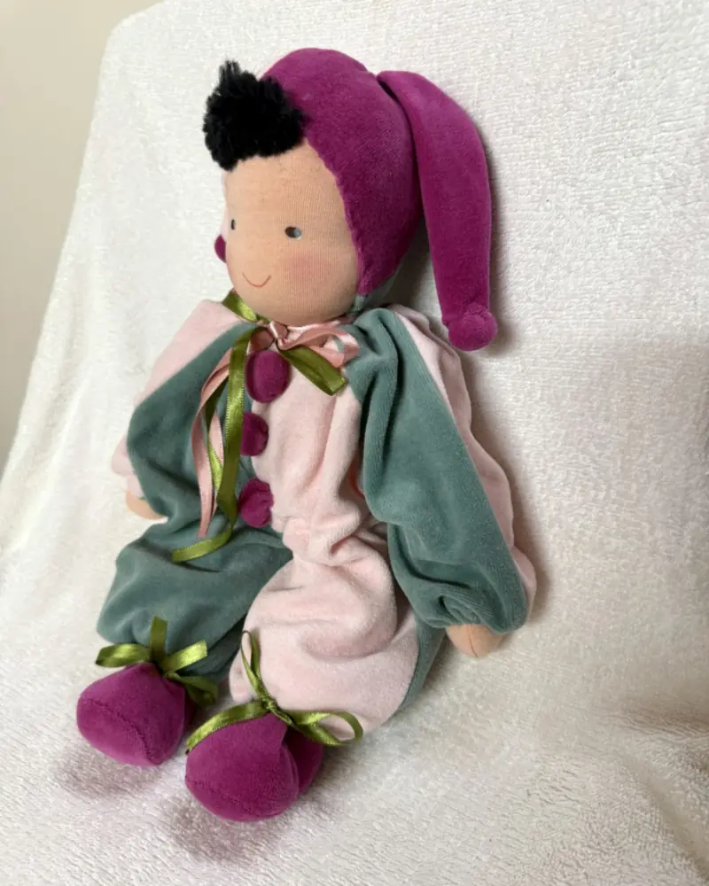 Polichinelo rosa e verde-acinzentado visto em ângulo, com o lado magenta do gorro e fitas de cetim verde e rosa