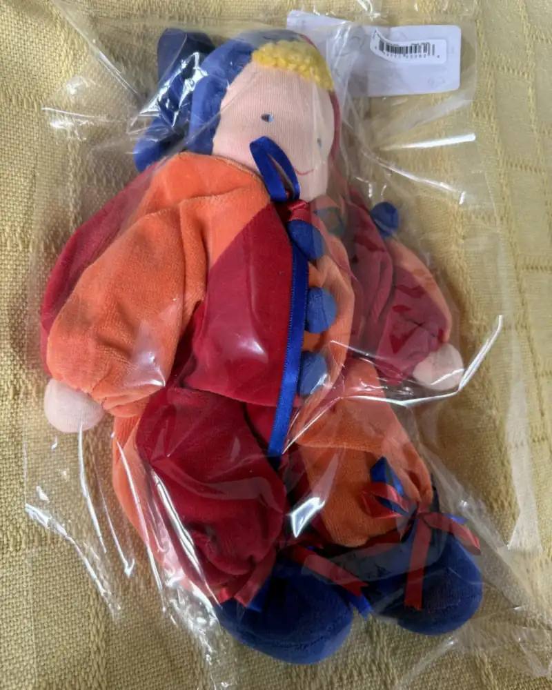 Polichinelo laranja e vermelho no saquinho de embalagem, com gorro azul, fita e botões-pompom azuis no peito