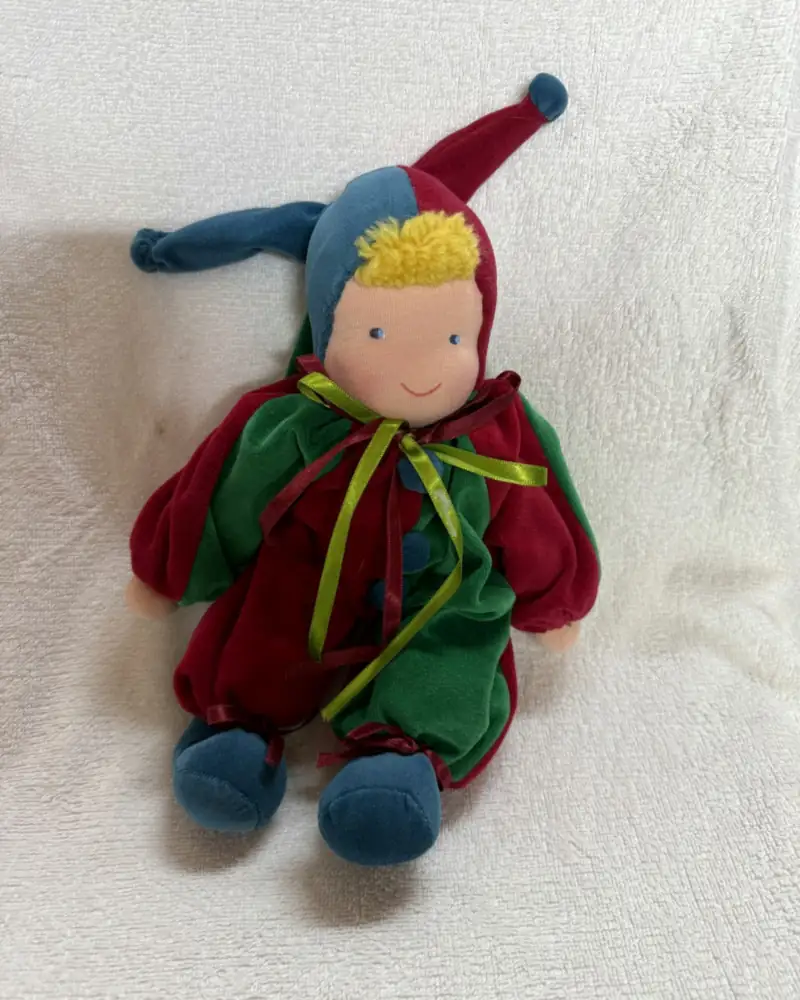 Polichinelo Pequeno Vermelho e Verde de pelúcia, de frente sobre manta branca, com gorro de duas pontas azul e vermelho
