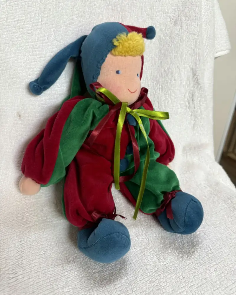 Polichinelo vermelho e verde visto em ângulo, com a ponta azul do gorro caída para o lado e botões-pompom azuis